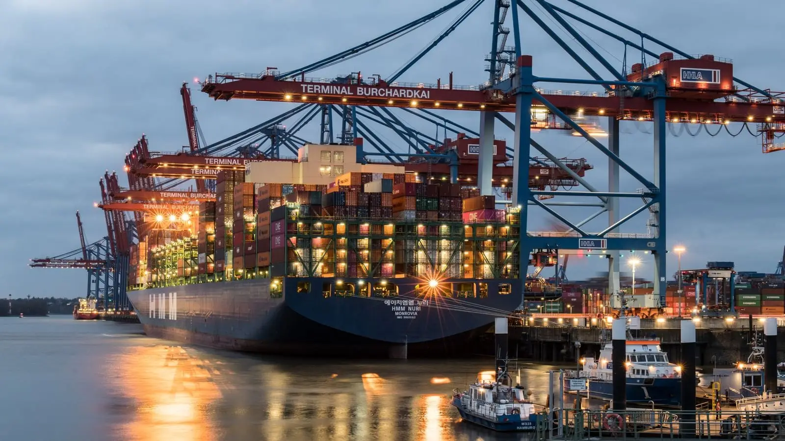 Container ships moored along a large terminal at dusk, cranes silhouetted above them
